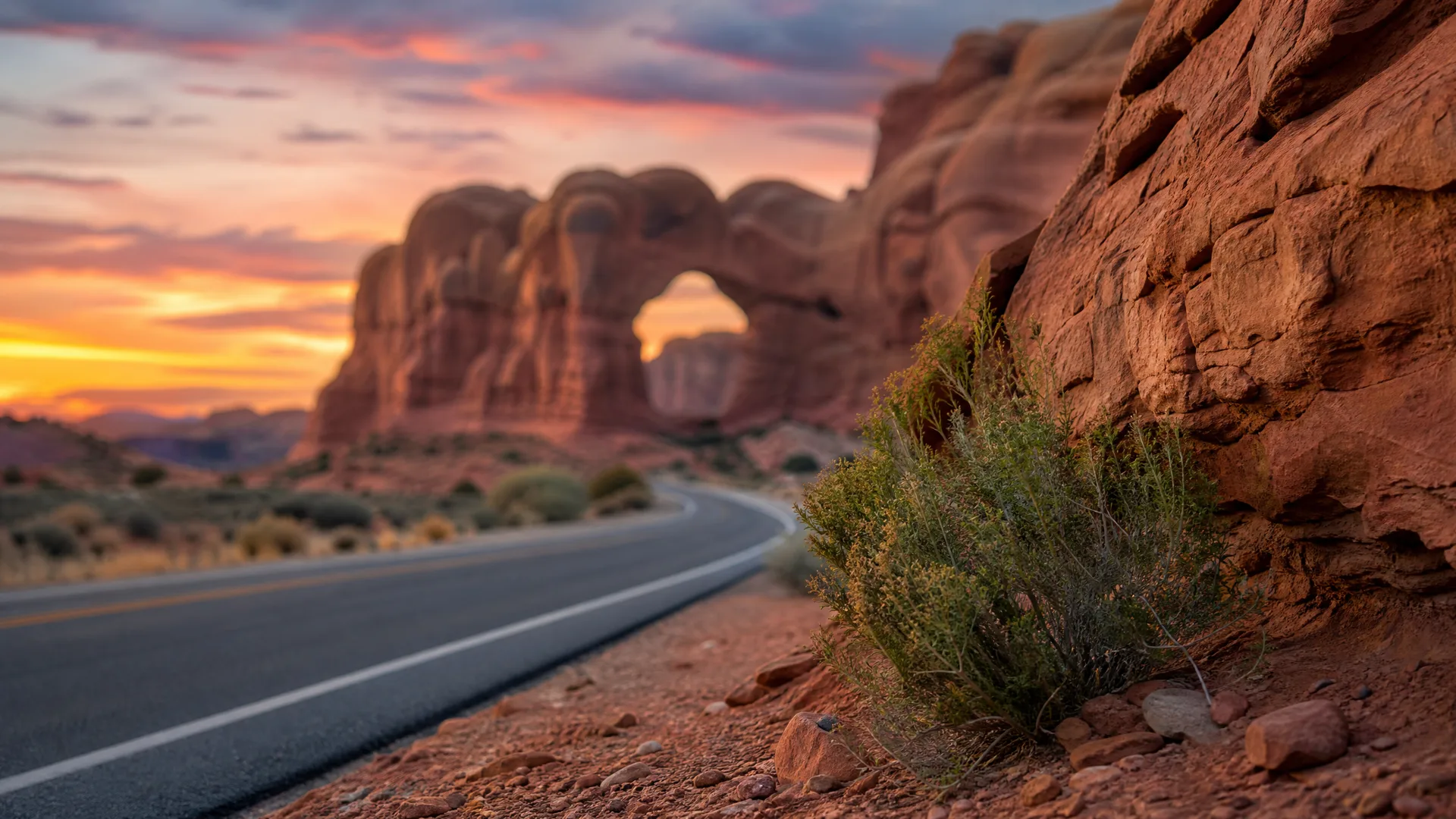 A two-lane paved road curves past red sandstone formations with a natural arch under a sunset sky, with a desert shrub and rocky ground in the foreground.